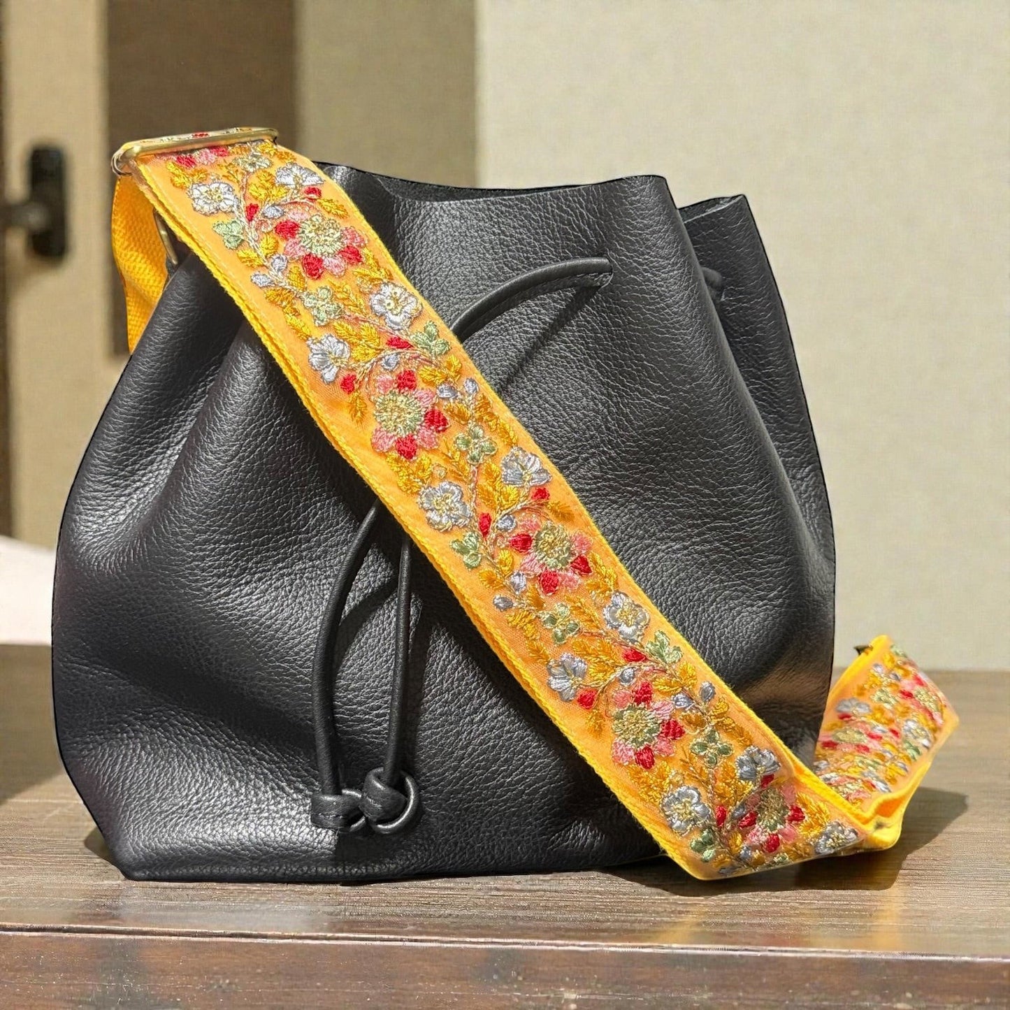 Rhea Artistry Studio yellow embroidered floral lace crossbody strap with brushed brass hardware on a black leather bucket bag.