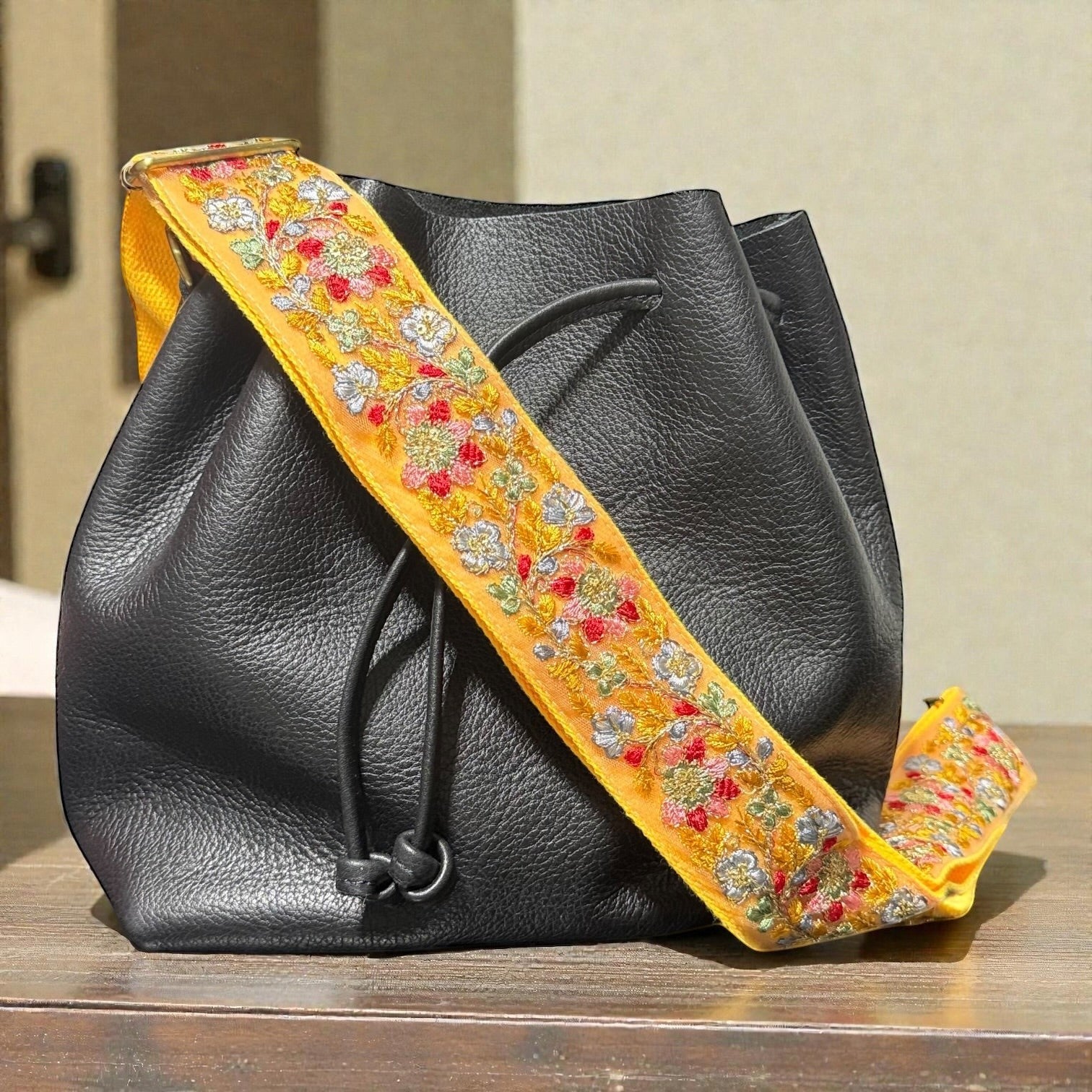 Rhea Artistry Studio yellow embroidered floral lace crossbody strap with brushed brass hardware on a black leather bucket bag.