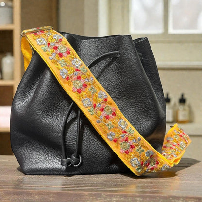 Rhea Artistry Studio yellow embroidered lace crossbody strap with floral details on a black leather bucket bag.