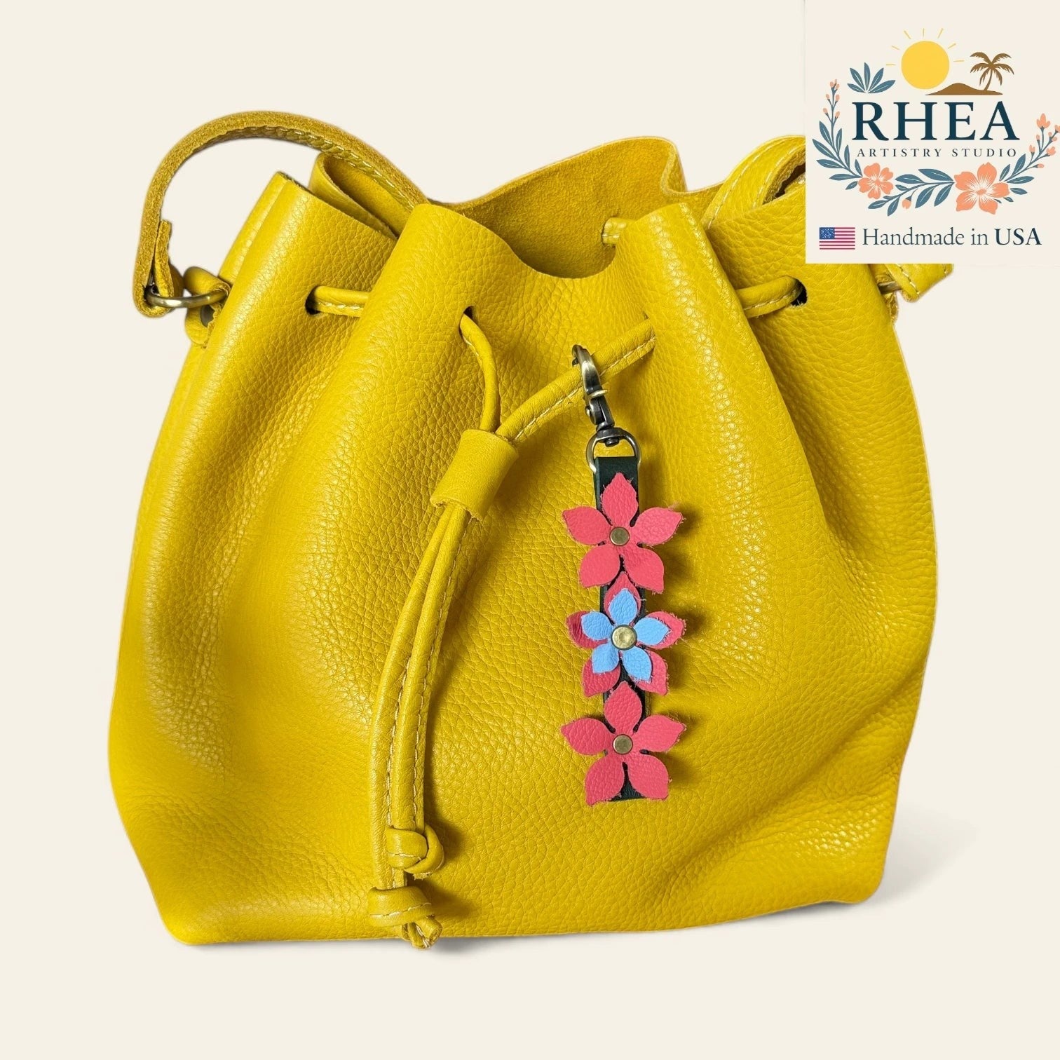 Rhea Artistry Studio yellow leather bucket bag with a floral charm featuring pink and blue leather flowers and a brushed brass clasp.