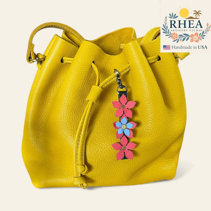 Rhea Artistry Studio yellow leather bucket bag with a floral charm featuring pink and blue leather flowers and a brushed brass clasp.