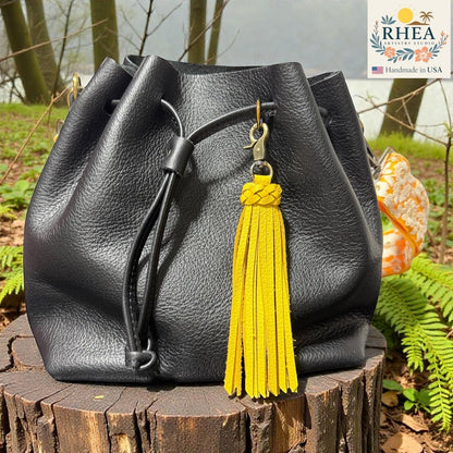 Rhea Artistry Studio yellow leather tassel bag charm with braided detail and brass clasp, attached to a black leather bucket bag.