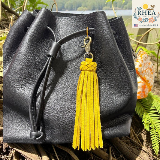 Rhea Artistry Studio yellow leather tassel bag charm with braided detail and brass clasp on a black leather bag.