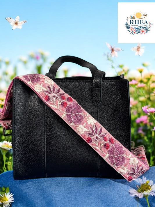 Black handcrafted leather handbag with a wide light pink strap embroidered in floral patterns of purple, magenta, and white, set against a wildflower field and blue sky — Rose Bloom Collection by Rhea Artistry Studio.