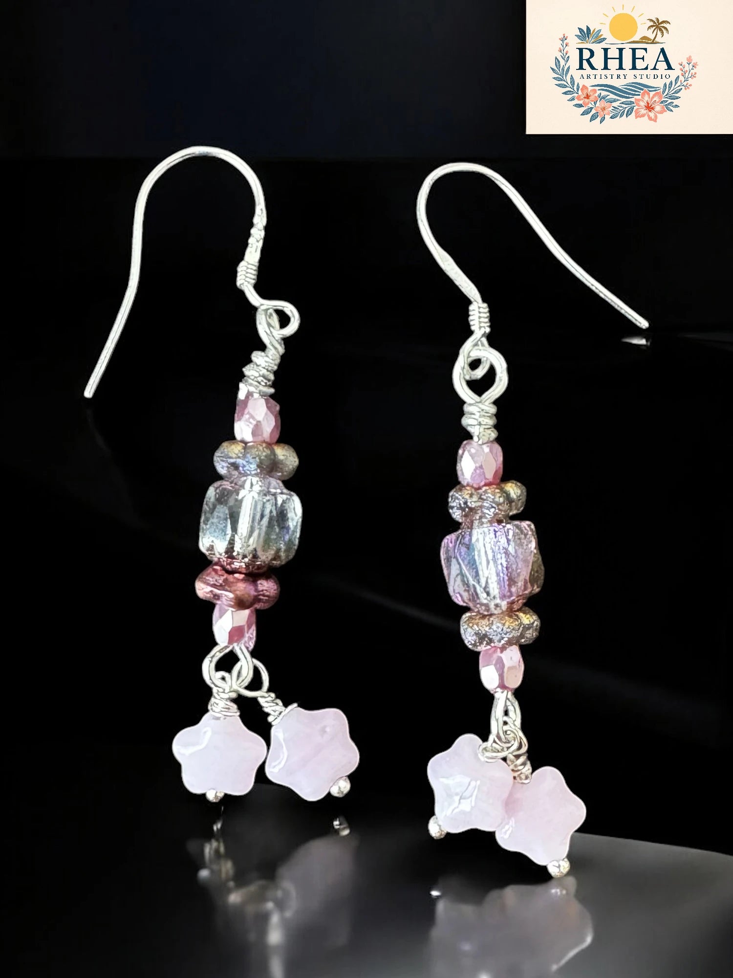 Pair of earrings with pink and clear beads on a dark background, branded 'RHEA'.
