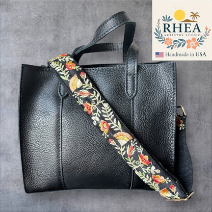 Black leather handbag with a floral purse strap on a gray background, featuring the Rhea Artistry Studio logo.