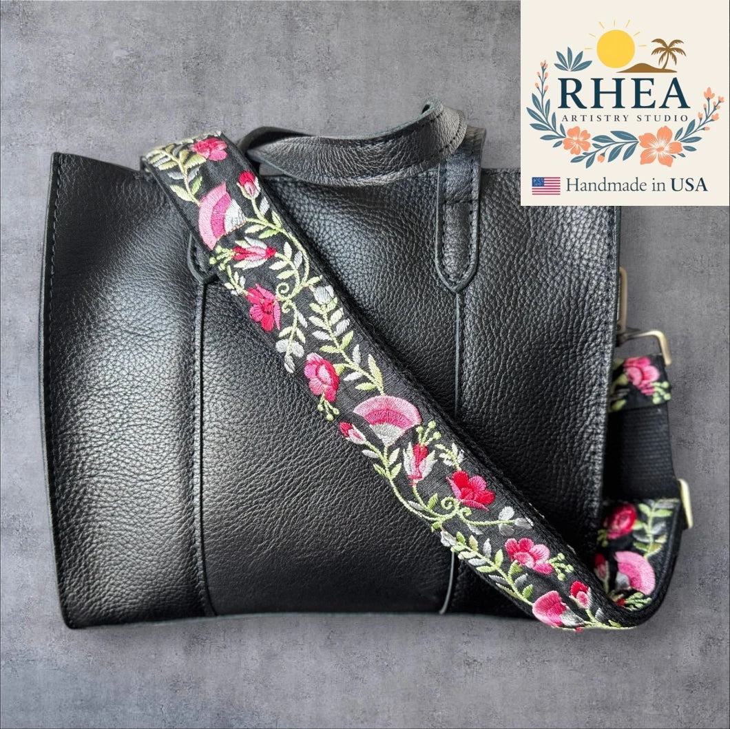Black leather handbag with a floral embroidered purse strap on a gray background, featuring the RHEEA Artistry Studio logo.