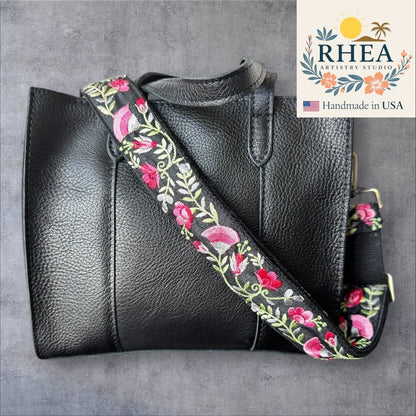 Black leather handbag with a floral embroidered purse strap on a gray background, featuring the RHEEA Artistry Studio logo.
