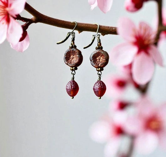 Earrings with red beads and gold accents hanging from a branch with pink flowers.