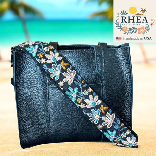 Black leather handbag with a floral embroidered strap on a beach background, featuring the RHEA Artistry Studio logo.