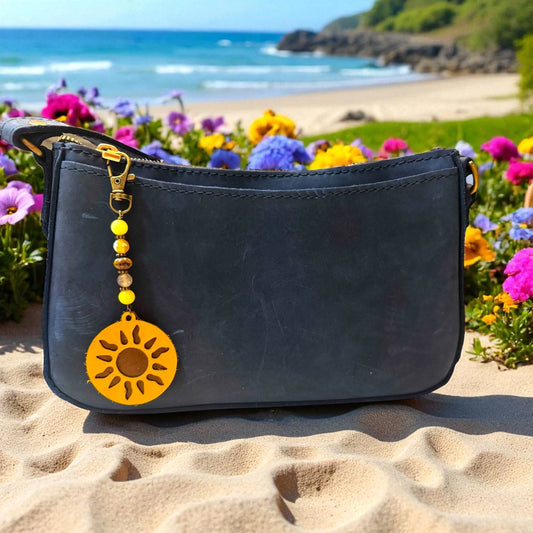 Black handbag with a yellow sun charm on a sandy beach with flowers and ocean in the background