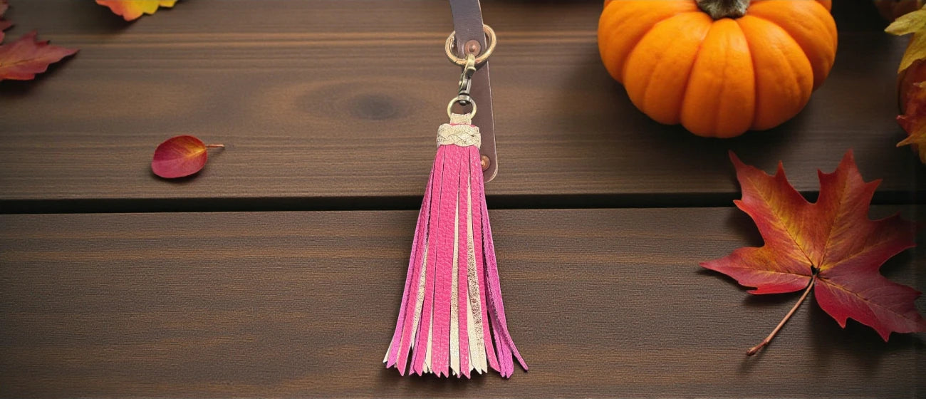 Pink tassel keychain on a wooden surface with autumn leaves and a pumpkin.