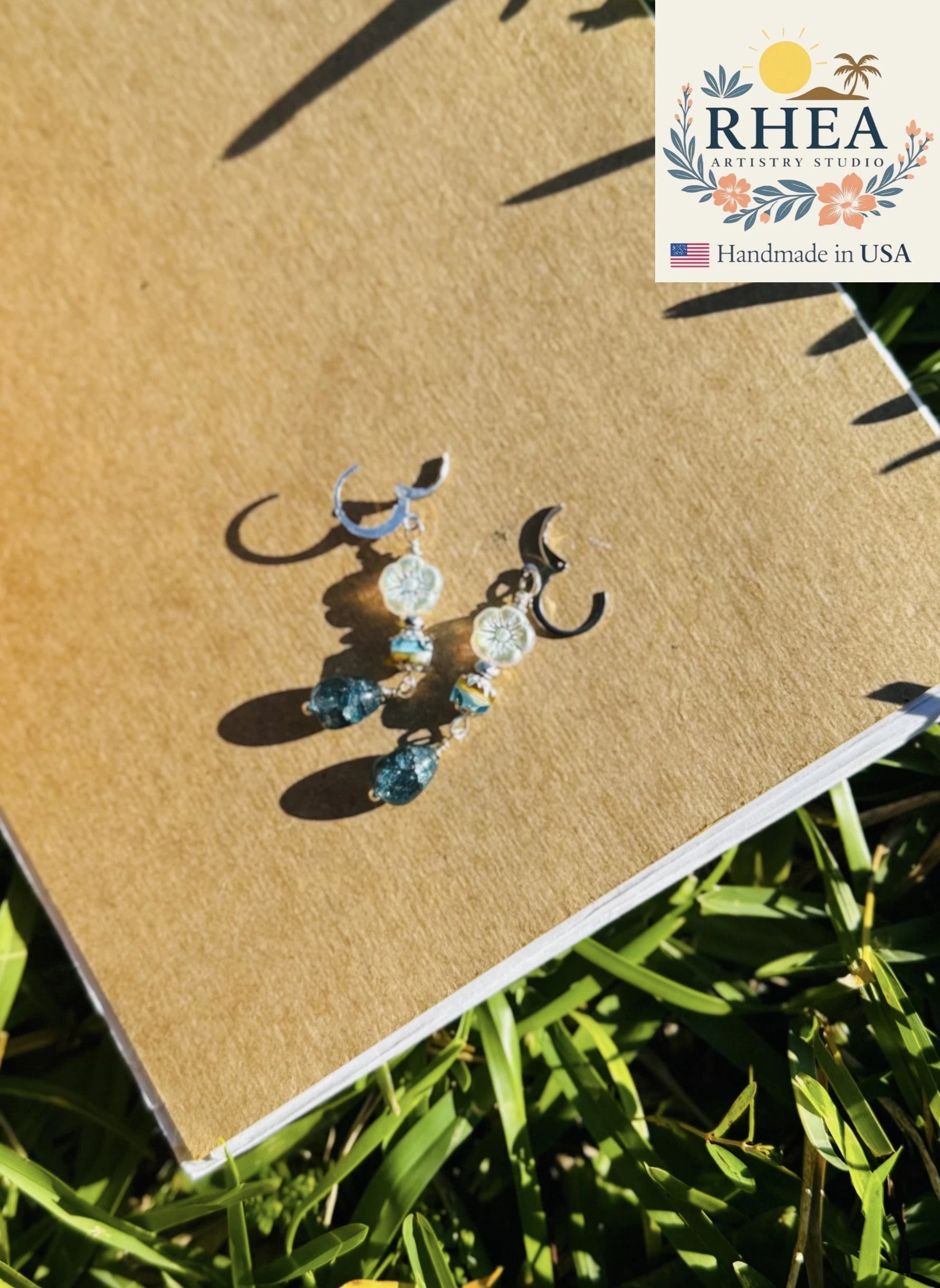 Earrings on a brown surface with 'RHEA' branding in the corner.