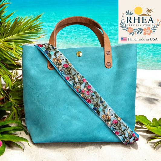 Turquoise tote bag with floral purse strap on a beach background, featuring the RHEA Artistry Studio logo.