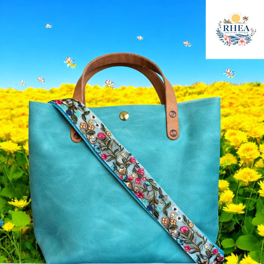 An aqua leather tote bag featuring the Tide Garden crossbody strap. The strap showcases botanical embroidery in vibrant greens, pinks, and mustard on a coastal blue base—photographed against a meadow setting.