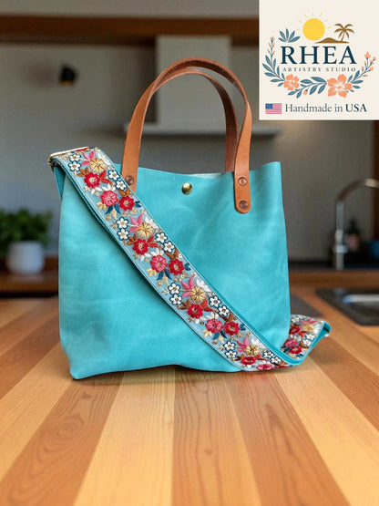 Turquoise handbag with floral strap on a wooden surface, RHEA brand visible
