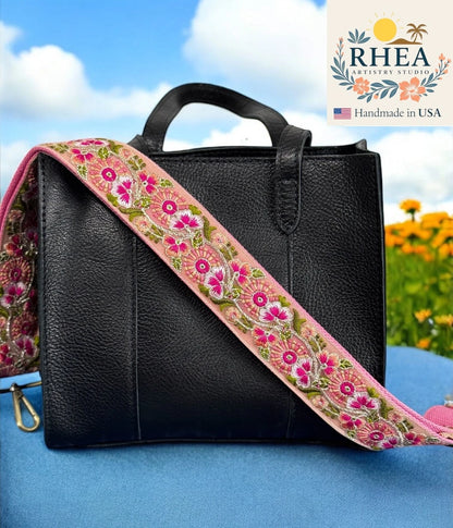 Black leather bag with a colorful floral purse strap against a blue sky and flower background, featuring the RHEA brand.