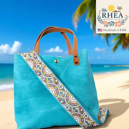 Turquoise tote bag with decorative purse strap on a beach, featuring RHEA branding.