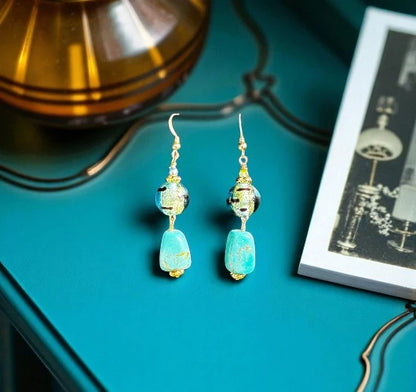 Pair of turquoise beaded earrings on a teal surface with a lamp and book in the background.