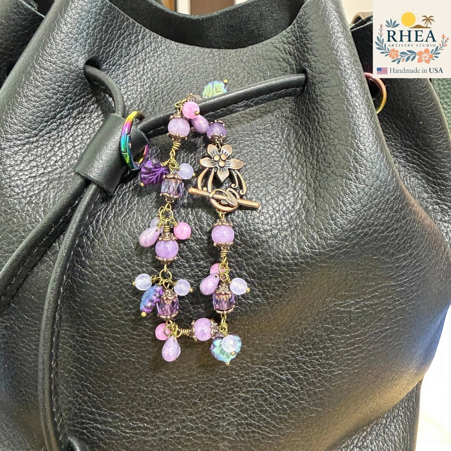Decorative purse charm with beads and flowers on a black leather bag, featuring the RHEA brand.