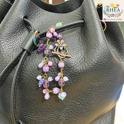 Decorative purse charm with beads and flowers on a black leather bag, featuring the RHEA brand.