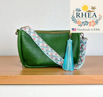 Green handbag with floral strap and tassel on a wooden surface, RHEA Artistry Studio logo visible.