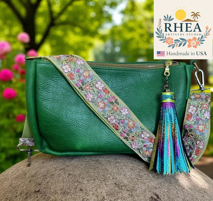 Green handbag with floral strap and tassel on a rock with blurred flowers and trees in the background, RHEA Artisan Studio logo visible.