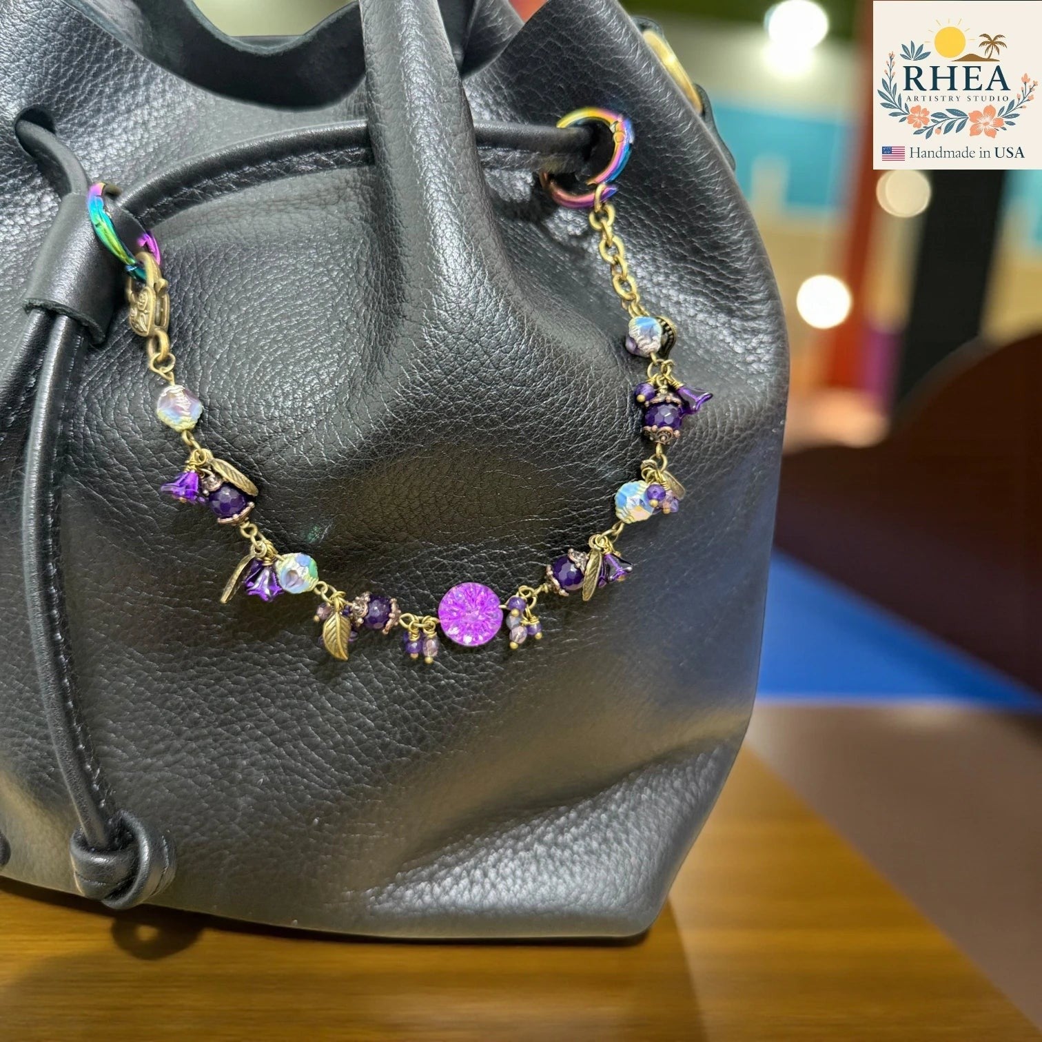 Black leather bag with a decorative bag charm featuring purple and gold elements on a blurred background.