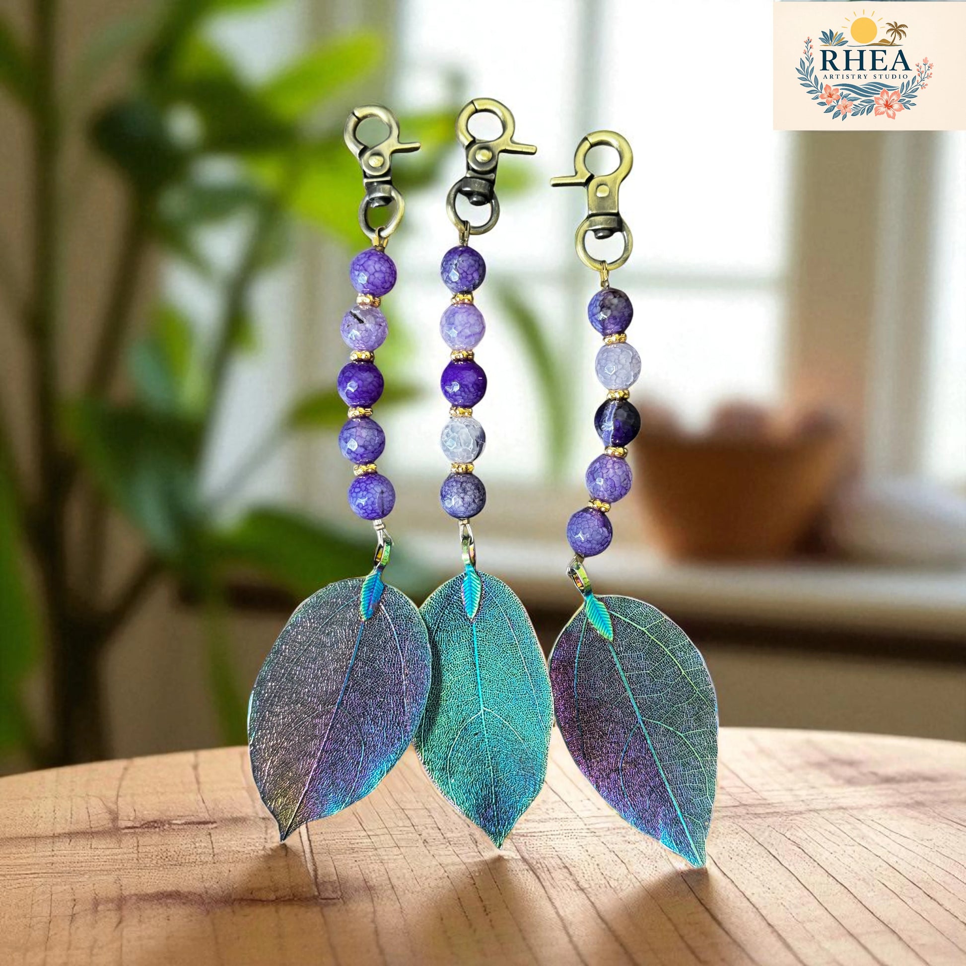 A handcrafted bag charms featuring multiple  single strands of violet-hued  gemstone beads and a shimmering electroplated leaf pendant, photographed on a minimal white marble background.