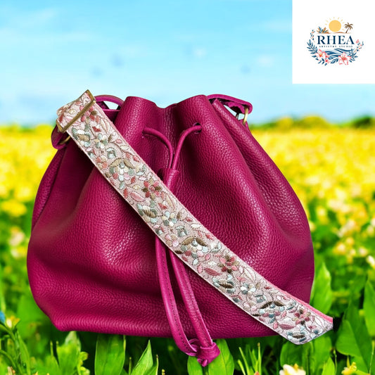 A romantic embroidered strap featuring soft pink and green vines with delicate white accents on a pastel base, attached to a magenta leather purse in the garden