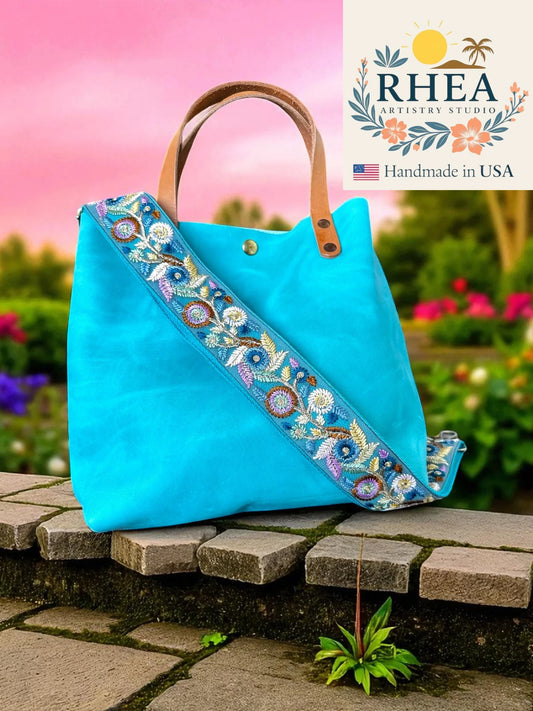 Blue tote bag with floral embroidery on a stone surface with a garden background, featuring the RHEA brand.