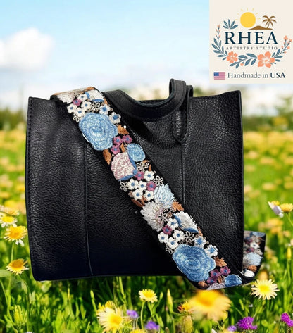 Black handbag with a decorative floral purse strap in a field of flowers, featuring the RHEA Artistry Studio logo.