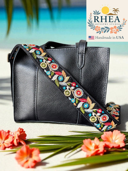 Black handbag with colorful floral strap on a beach setting, featuring the RHEA brand.