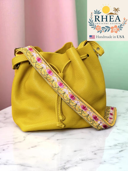Yellow handbag with floral purse strap on a marble surface, RHEA brand visible.