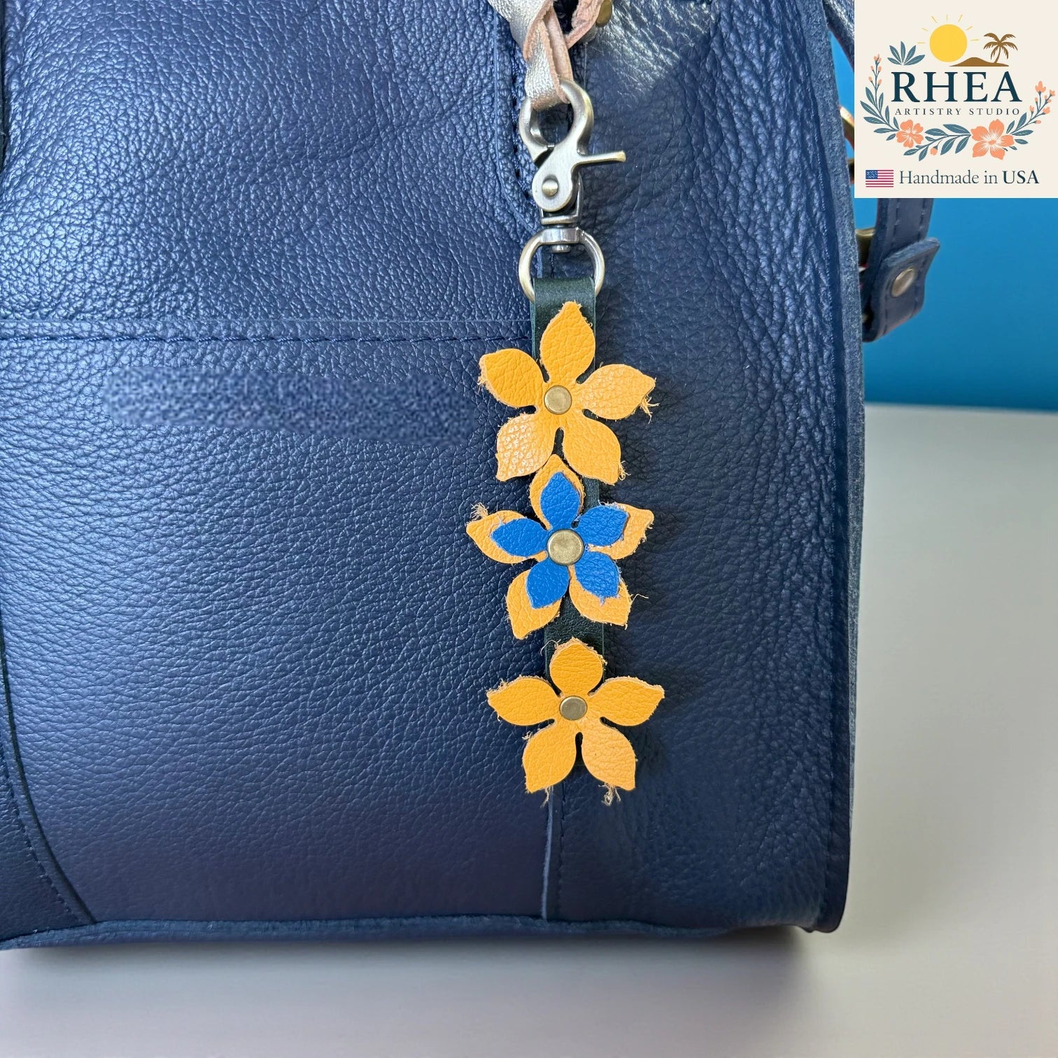 Blue leather bag with yellow and blue floral keychain charm on a white surface, RHEA brand visible.