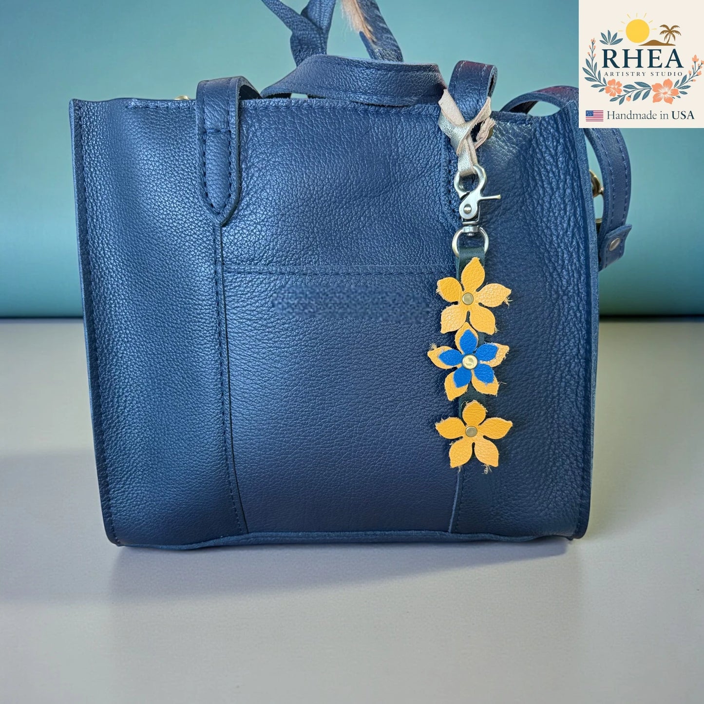 Blue handbag with a floral keychain charm on a light gray surface, branded 'RHEA'.