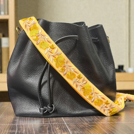 Embroidered shoulder strap in a 1.5 inch width, Black leather handbag with a decorative yellow strap on a wooden surface.