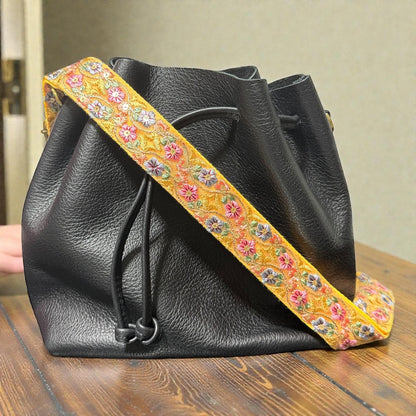 Cotton webbing backing on a 1.5 inch embroidered lace strap, Black leather drawstring bag with a colorful floral strap on a wooden surface.