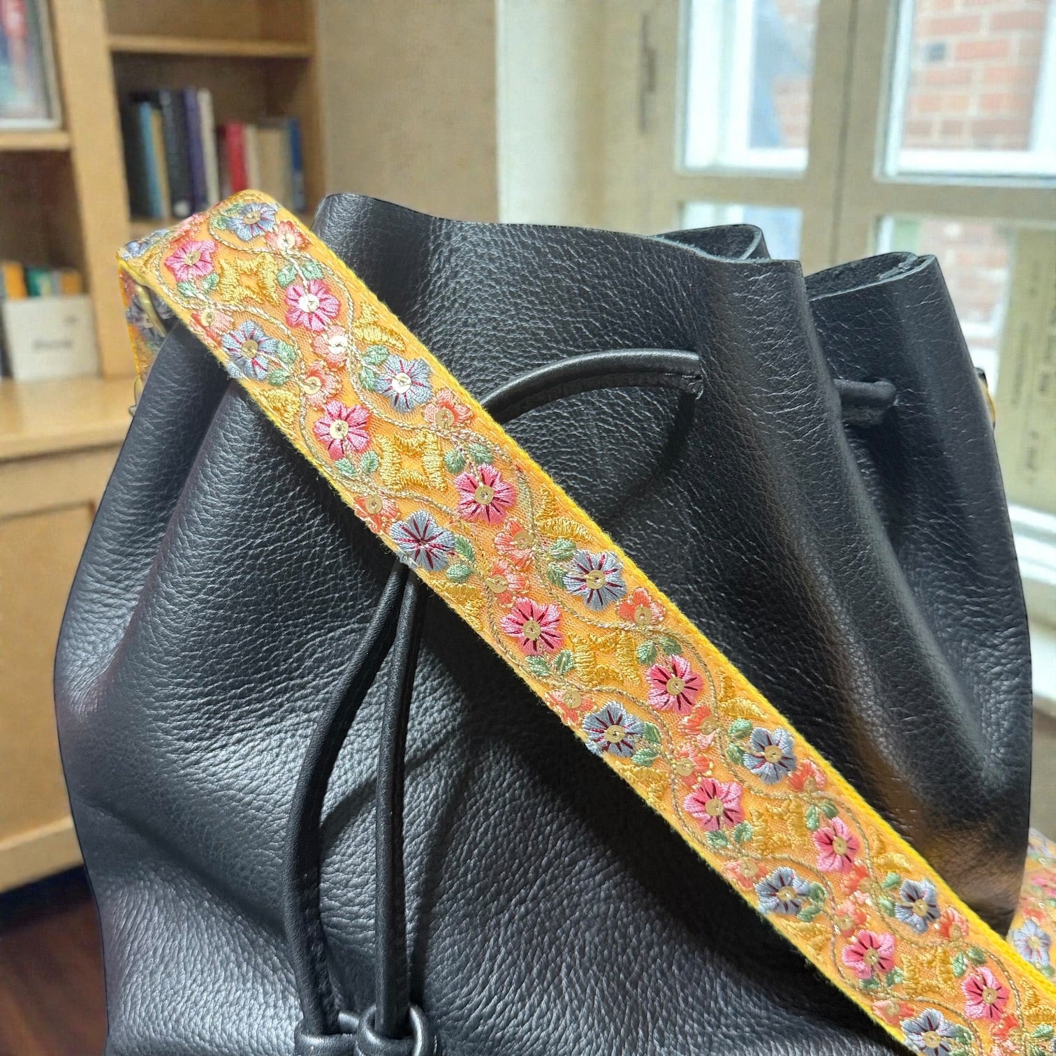 Pattern detail of an embroidered strap in a 1.5 inch width, Black leather bag with a colorful floral strap in an indoor setting