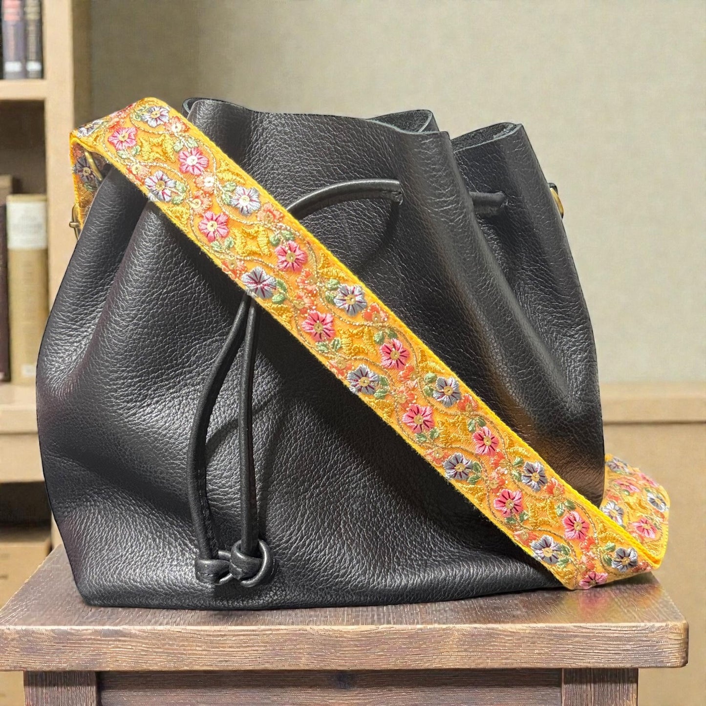  Embroidered crossbody strap in a 1.5 inch width, Black leather bag with a colorful floral strap on a wooden surface