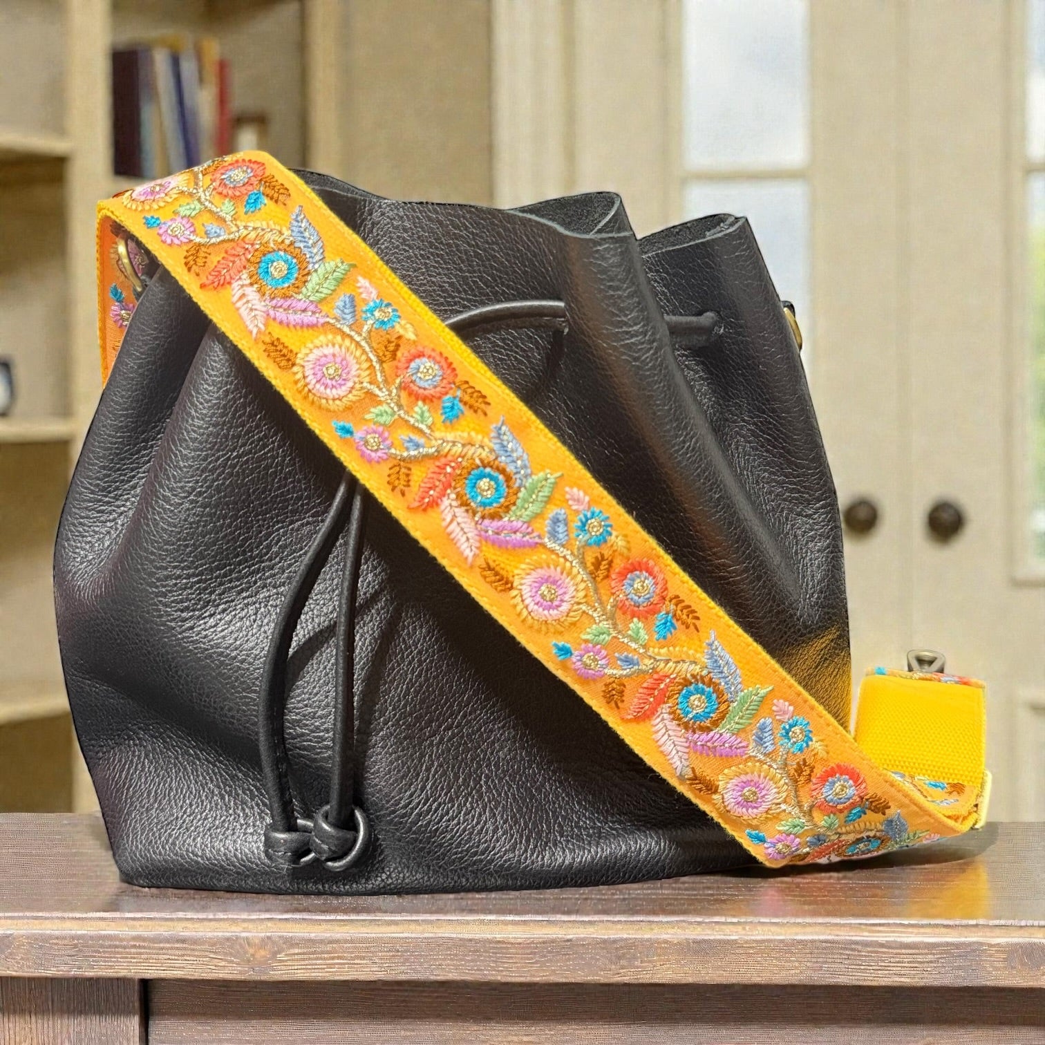 Floral embroidered strap attached to a handbag, Black leather bag with a colorful embroidered strap on a wooden surface.