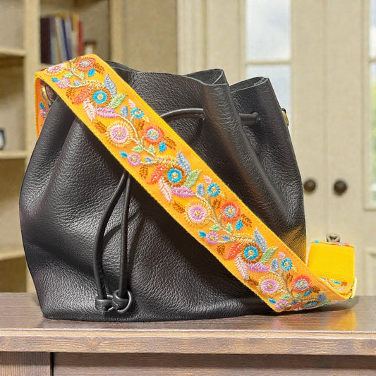 Floral embroidered strap attached to a handbag, Black leather bag with a colorful embroidered strap on a wooden surface.