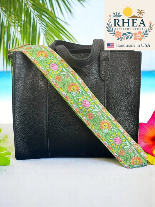 Black leather bag with a colorful floral purse strap against a tropical background, featuring the RHEA brand.