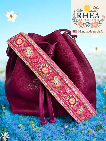 Pink leather bag with a floral purse strap on a blue floral background, featuring the RHEA Antiquities Studio logo.